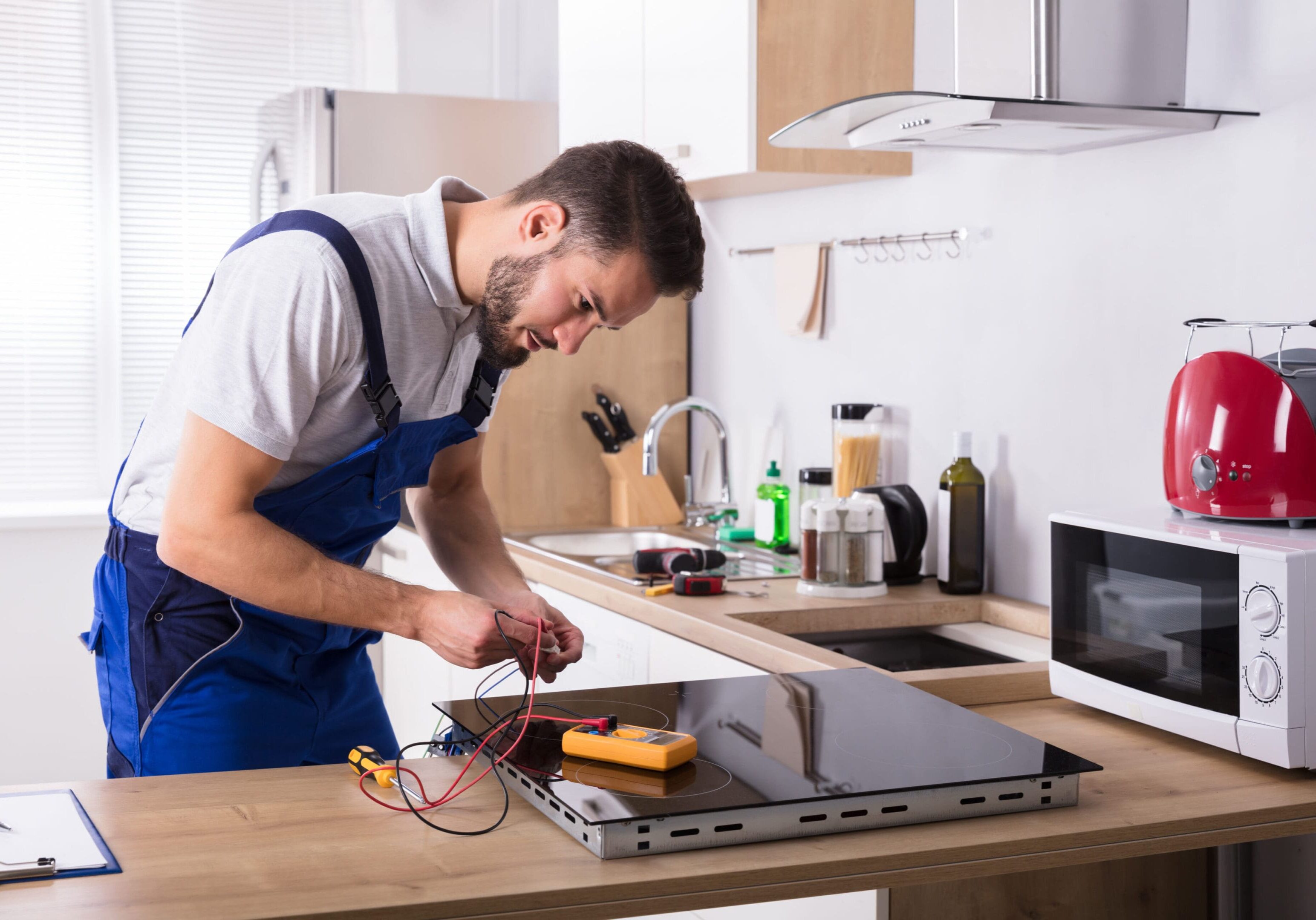 Male Technician Repairing Induction Stove With Digital Multimeter In Kitchen