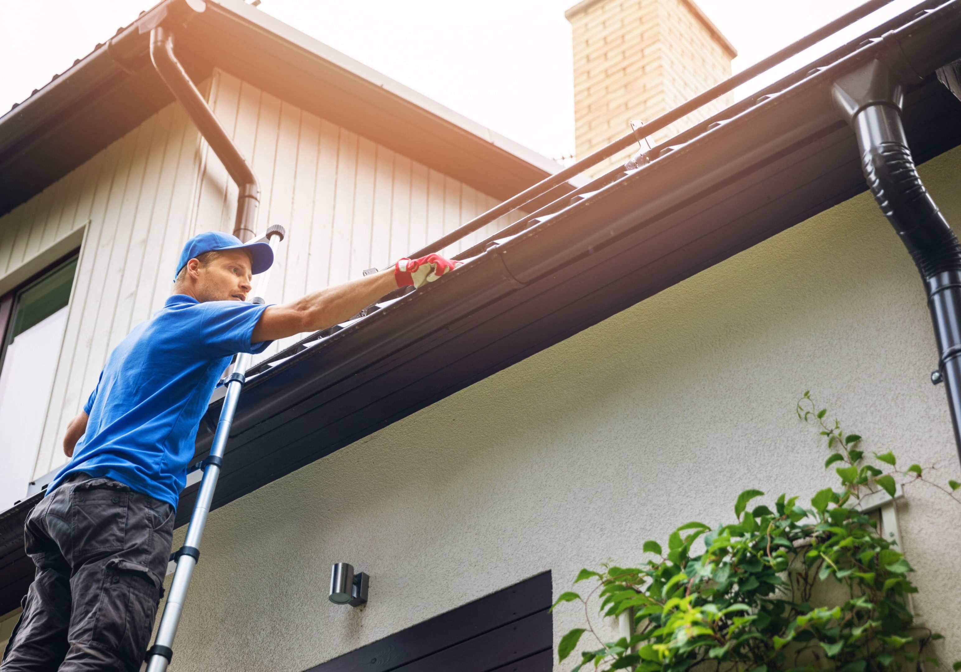 man on ladder cleaning house gutter from leaves and dirt